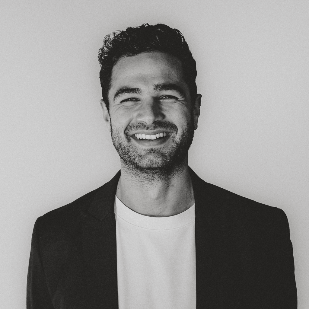 Schwarzwei&szlig;-Portr&auml;t eines l&auml;chelnden Mannes im Sakko und T-Shirt.